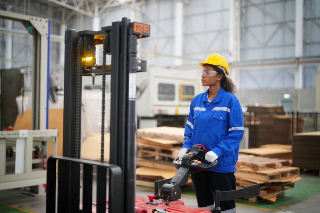 A woman in a blue shirt and hard hat standing next to a forklift